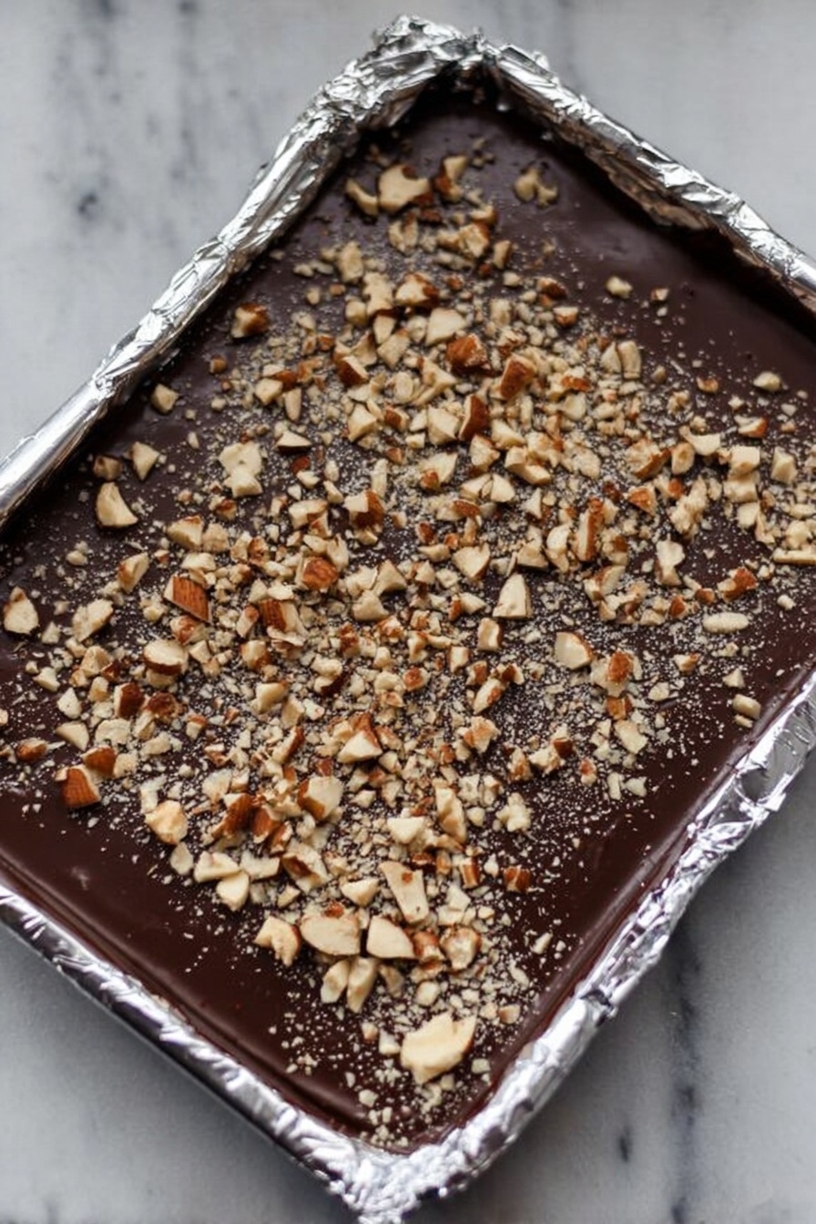 English Toffee with Chocolate and Nuts, Easy Toffee Recipe, Homemade Toffee, Crunchy Toffee Treats, Chocolate Nut Toffee - This image shows a foil-lined rectangular tray filled with a smooth, dark chocolate layer. On top of the chocolate is an even spread of chopped nuts, both larger and smaller pieces, creating a textured surface. The foil edges around the tray are crinkled and shiny, reflecting light softly. The background is a white marbled texture. photo taken with an iphone --ar 2:3 --v 7