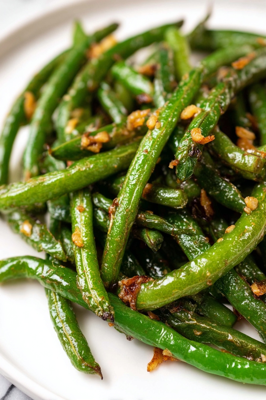 Garlic Green Beans Stir Fry, green beans stir fry, healthy vegetable recipes, quick garlic green beans, savory green bean side dish - A pile of cooked green beans with browned, slightly wrinkled skin is placed on a white paper towel over a white marbled surface. To the upper left of the green beans, there is a clear round glass bowl filled with a light brown sauce mixed with small pieces of chopped garlic or onion. The sauce has a glossy texture. The background is a white cloth that partially covers the surface. photo taken with an iphone --ar 2:3 --v 7