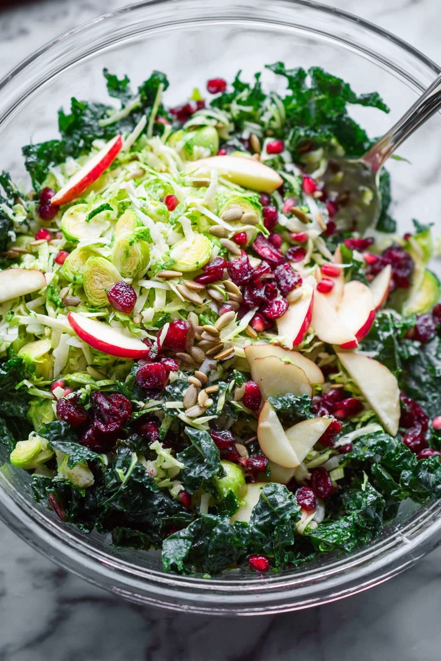 Shaved Brussels Sprouts Salad with Pomegranate, Brussels Sprouts Salad, Healthy Pomegranate Salad, Vibrant Vegetables Salad, Easy Fall Salad - A clear glass bowl filled with a fresh salad on a white marbled surface, showing multiple layers of ingredients. The base layer includes dark green leafy kale with a thick texture. On top, there is a layer of thinly sliced light green Brussels sprouts mixed with small slices of light red and white apple. Small pieces of red onion add specks of purple color throughout. Scattered across the salad are bright red dried cranberries and small shiny red pomegranate seeds. Light brown sunflower seeds are spread over the salad, adding texture. A silver serving spoon is partially visible inside the bowl. The whole scene is bright and fresh, with natural light highlighting the crispness of the vegetables and fruits. Photo taken with an iphone --ar 2:3 --v 7