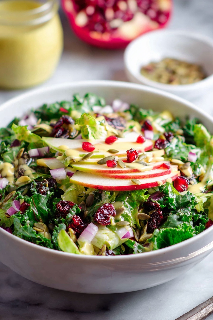 Shaved Brussels Sprouts Salad with Pomegranate, Brussels Sprouts Salad, Healthy Pomegranate Salad, Vibrant Vegetables Salad, Easy Fall Salad - A white bowl is filled with a fresh salad showing multiple layers: at the bottom are bright green leafy vegetables, followed by thin slices of red and green apple. On top, there are pieces of chopped red onion, dark green kale leaves, bright red pomegranate seeds, dried red cranberries, and scattered sunflower seeds, all mixed together. The salad is lightly dressed with a pale yellow dressing that adds a slight shine to the ingredients. The bowl is placed on a white marbled surface, with a blurred jar of light yellow dressing and a small white bowl of seeds in the background. photo taken with an iphone --ar 2:3 --v 7
