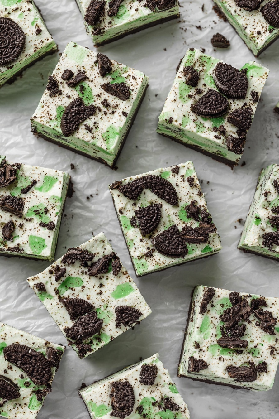 Mint Oreo White Chocolate Fudge, mint Oreo fudge, white chocolate fudge recipes, no-bake fudge desserts, creamy mint fudge - The image shows many square pieces of dessert laid out on crumpled white paper over a white marbled surface. Each square has two visible layers: the bottom layer is a light creamy color with specks inside, and the top layer is a white cream with crushed dark chocolate cookies that have bright green filling scattered unevenly across the surface. The cookies add a rough texture on top, with pieces varying in size and shape. The squares are arranged closely but not touching each other, filling the image space evenly, and giving a look of a cool, creamy treat with a minty cookie crunch on top photo taken with an iphone --ar 2:3 --v 7