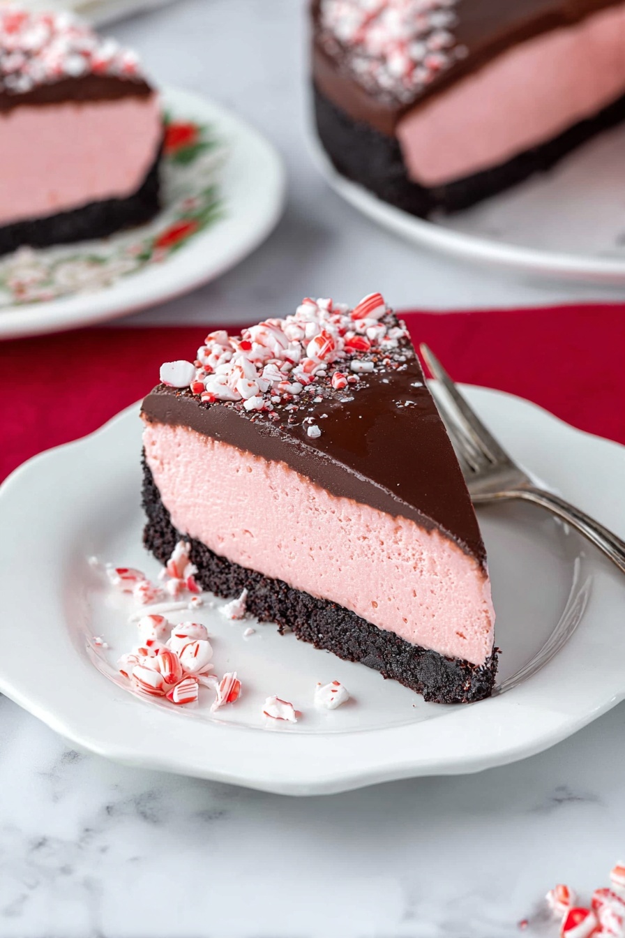 Peppermint Cheesecake with Chocolate Ganache, peppermint cheesecake, holiday cheesecake, festive dessert recipes, easy cheesecakes - A slice of dessert with three distinct layers sits on a white plate with scalloped edges; the bottom layer is a dark, crumbly crust, the middle layer is a thick, smooth pink mousse, and the top layer is a glossy dark chocolate coating sprinkled with crushed red-and-white candy pieces. The slice is garnished with more crushed candies at the base and is accompanied by a silver fork placed nearby. In the background, parts of the full dessert and another slice on a white plate with floral patterns rest on a white marbled surface, accented by a red cloth. Photo taken with an iphone --ar 2:3 --v 7
