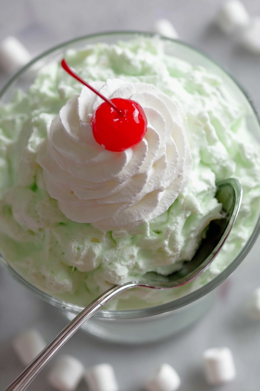 Watergate Salad Pineapple Marshmallows, Watergate Salad Recipe with Pineapple, Easy Watergate Salad, No-Bake Fruit Salad, Retro Dessert Recipes - A close-up image of a layered dessert in a clear glass bowl placed on a white marbled surface with mini white marshmallows scattered around. The bottom layer is a creamy, light green textured mixture that looks fluffy and thick. On top of this green layer is a smooth, white dollop of whipped cream shaped in a spiral pattern. Sitting right in the center of the whipped cream is a bright red, shiny cherry. A silver spoon is partly inserted into the green layer, curving around the side of the bowl. photo taken with an iphone --ar 2:3 --v 7