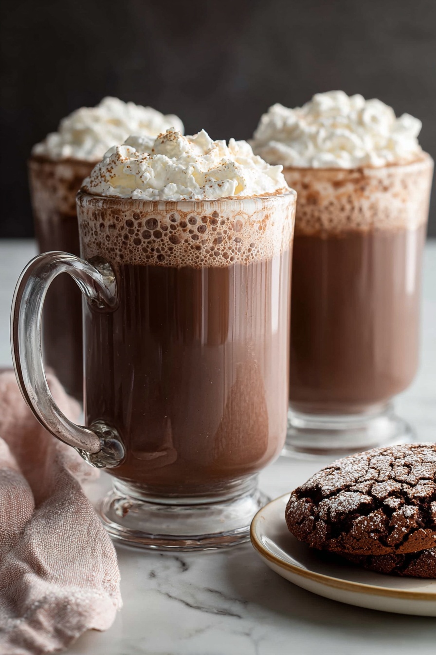 Creamy Homemade Hot Chocolate, best hot chocolate, homemade hot chocolate recipe, rich hot cocoa, comfort drink recipes - Two clear glass mugs filled with rich brown hot chocolate, topped with a thick layer of light white whipped cream that has a fluffy texture and soft peaks. The mugs have sturdy handles positioned on the left, and the hot chocolate shows slight bubbles near the edges under the whipped cream. In the foreground, on the right side, there is a white plate with a beige rim holding two cracked chocolate cookies with a powdery surface. A soft pink and white cloth napkin is casually placed on the left side near the front mug. All items sit on a smooth white marbled surface with a dark, blurred background behind them. Photo taken with an iphone --ar 2:3 --v 7