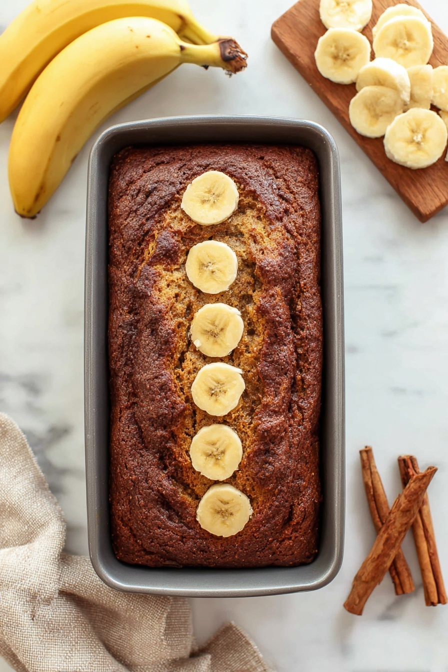 Cinnamon Swirl Banana Bread, banana bread with cinnamon swirl, moist banana bread recipe, quick banana bread, easy cinnamon swirl recipe - A thick rectangular loaf of dark brown banana bread rests in a grey baking pan, with five round banana slices lined up in the center top of the bread. To the left, two whole yellow bananas lay curved on a white marbled surface. To the right, a wooden board holds several light yellow banana slices arranged in overlapping circles. At the bottom right corner, three brown cinnamon sticks rest on the white marbled surface, and a beige cloth is partially visible at the bottom left corner. photo taken with an iphone --ar 2:3 --v 7