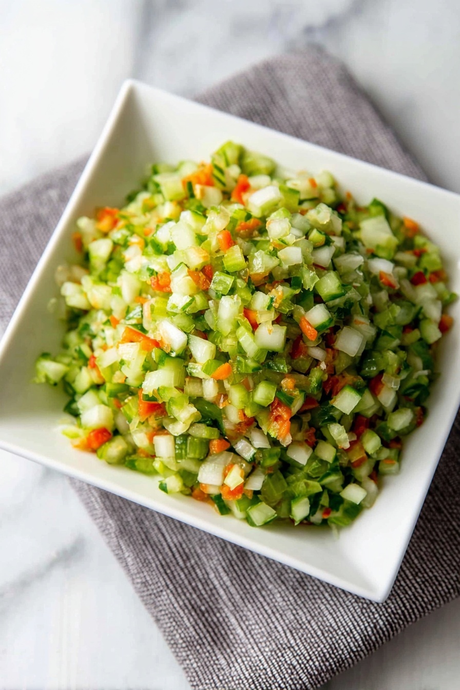 Dill Pickle Salsa, Dill Pickle Salsa recipe, tangy pickle salsa, crunchy pickle salsa, quick salsa recipe - A close-up view of finely chopped vegetables in a white square bowl placed on a grey cloth on a white marbled surface. The mix has small pieces with mostly green colors from celery or cucumber, bright orange bits from carrots, and some white pieces from onions, creating a fresh and colorful look. The texture appears crisp and moist, with the small vegetable pieces piled up in layers that give a sense of volume and freshness. photo taken with an iphone --ar 2:3 --v 7