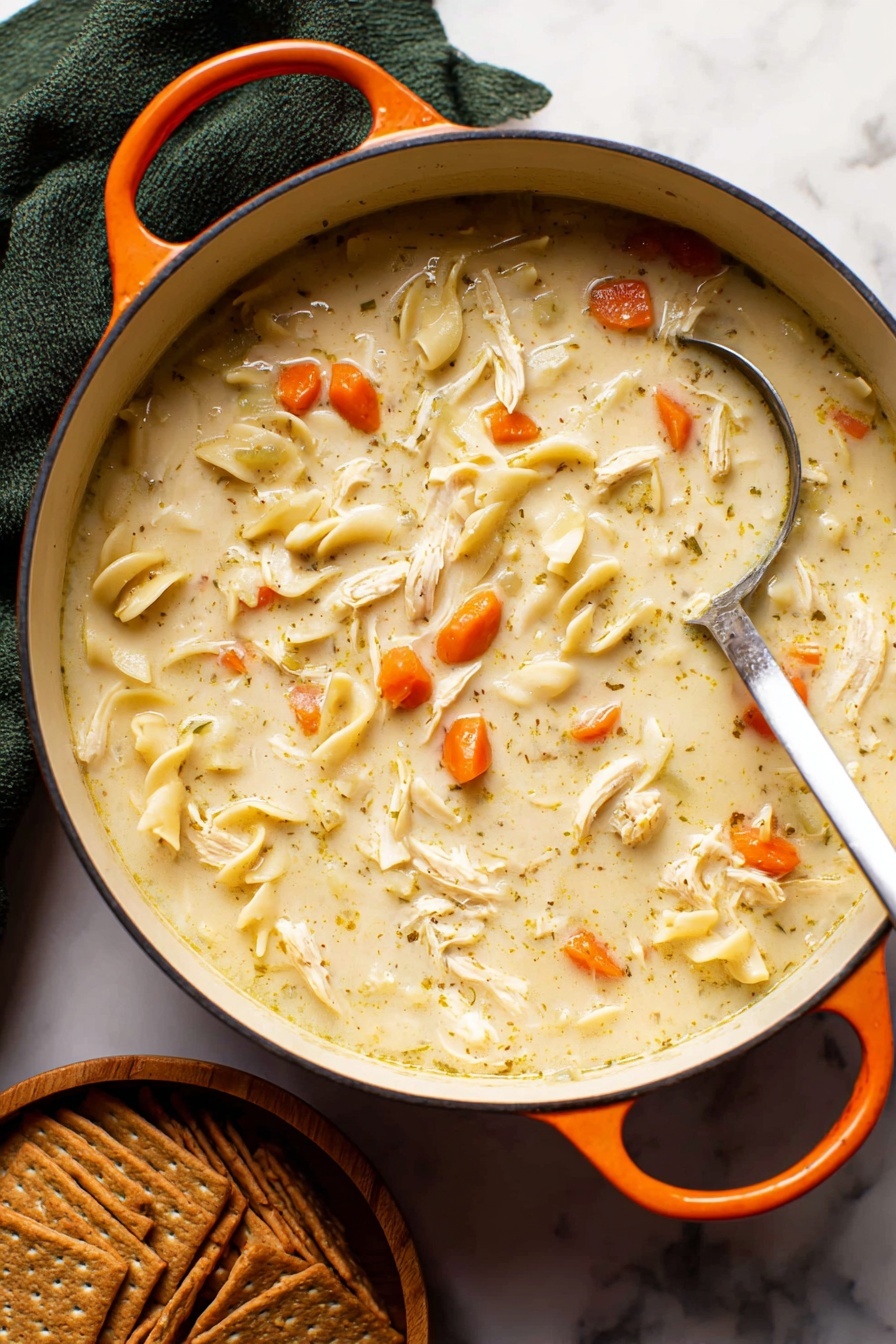 Creamy Chicken Noodle Soup, comfort food recipes, easy chicken soup, hearty noodle soup, homemade chicken noodle soup - A close-up of a white pot filled with creamy chicken noodle soup, showing a thick light beige broth mixed with small bright orange carrot slices, soft shredded white chicken pieces, and flat light yellowish noodles scattered throughout. A silver ladle is partially dipped in the soup, resting on the right side of the pot. The pot has an orange handle on each side and sits on a white marbled surface. Below the pot, there is a stack of brown crackers arranged neatly in a small wooden bowl. A dark green cloth is folded behind the pot, adding contrast to the scene. Photo taken with an iphone --ar 2:3 --v 7