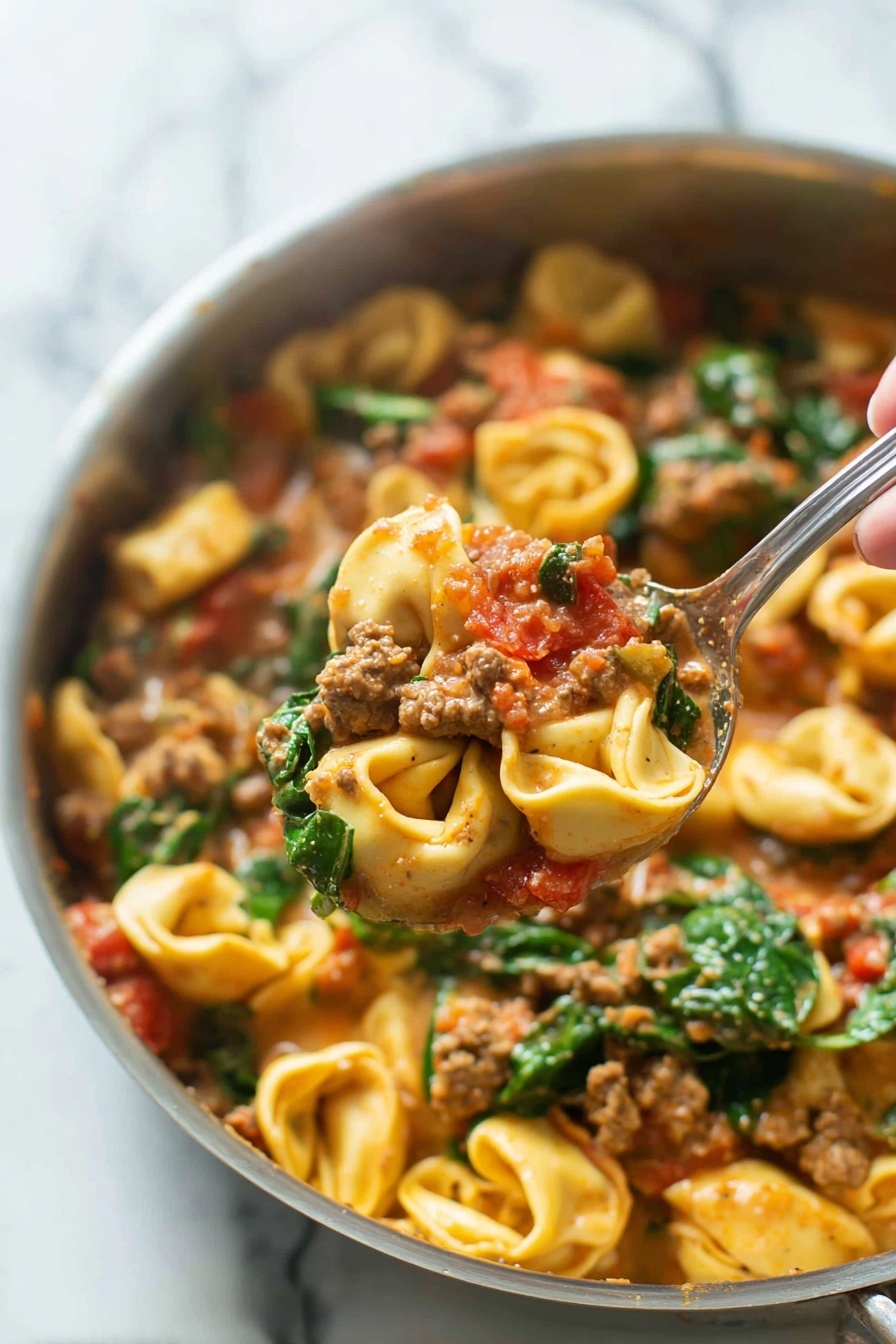 One-Pan Sausage Tortellini, easy sausage tortellini dinner, quick sausage tortellini meal, creamy tortellini skillet, hearty sausage pasta - A close-up image of a cooked pasta dish in a silver pan shows three main layers. The bottom layer is pasta tortellini, small ring-shaped with light yellow color and slightly ruffled edges. The middle layer features ground meat chunks mixed with pieces of red tomato, all coated in a creamy, light brown sauce. The top layer has fresh green spinach leaves scattered throughout, adding vibrant color and texture. A woman's hand holding a silver spoon lifts a portion of the pasta, meat, tomato, and spinach mixture above the pan. The background is a white marbled surface. photo taken with an iphone --ar 2:3 --v 7