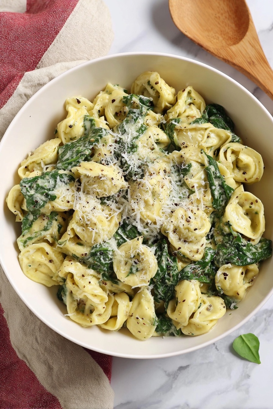 Creamy Pesto Tortellini with Spinach, easy pesto tortellini, quick weeknight pasta dinner, vegetarian pasta recipes, flavorful cheese tortellini - A white bowl filled with three layers of tortellini pasta that is light yellow, mixed with dark green spinach leaves, and coated in a creamy sauce with flecks of black pepper and herbs. On top, there is a light sprinkle of grated white cheese finely covering some pasta and spinach. The bowl sits on a white marbled surface with a beige and red cloth folded beside it, and a wooden spoon rests nearby, all softly lit in natural light. Photo taken with an iphone --ar 2:3 --v 7