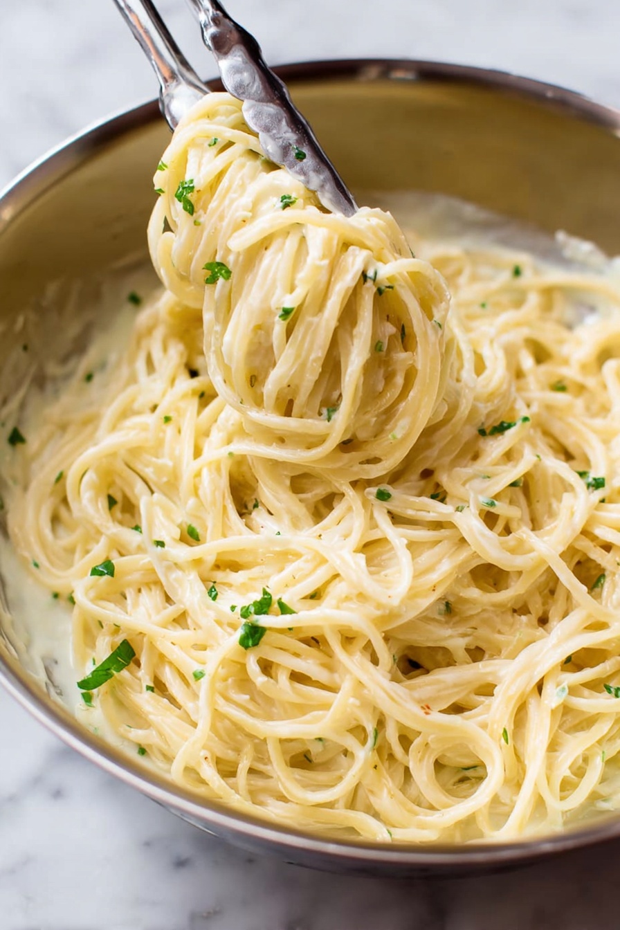 Garlic Parmesan Pasta, easy garlic pasta recipe, creamy garlic pasta, one-pot garlic pasta, quick Parmesan pasta - A close-up view of a stainless steel bowl filled with creamy spaghetti pasta covered in a smooth, pale yellow sauce. The pasta strands are thick and smoothly coated, twirled around a pair of metal tongs that are lifting some of the noodles from the bowl. Small bits of green herbs are sprinkled lightly over the pasta, adding a touch of color contrast. The background is a white marbled texture. photo taken with an iphone --ar 2:3 --v 7