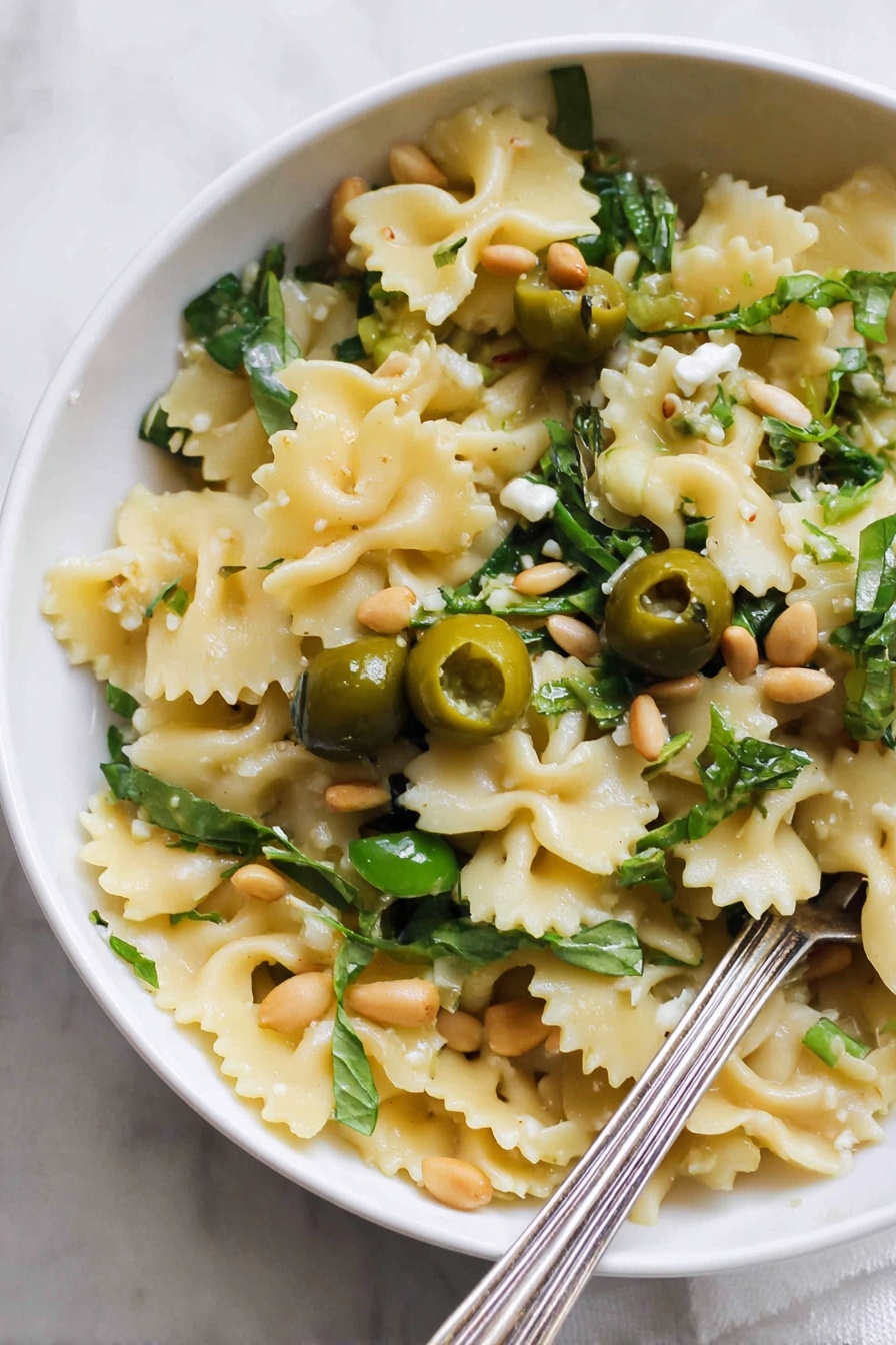 Dirty Martini Pasta Salad, pasta salad with olives and lemon, savory pasta salad recipe, summer pasta salad ideas, bold flavored pasta salad - A close-up view of a white bowl filled with one layer of wavy-edged pasta pieces that are light yellow in color. Scattered on top are halved green olives, fresh green leafy herbs, and a few light brown pine nuts. There are also small pieces of a creamy white ingredient and some garlic cloves. A silver fork rests inside the bowl at the bottom right corner. The bowl is placed on a white marbled surface. Photo taken with an iphone --ar 2:3 --v 7