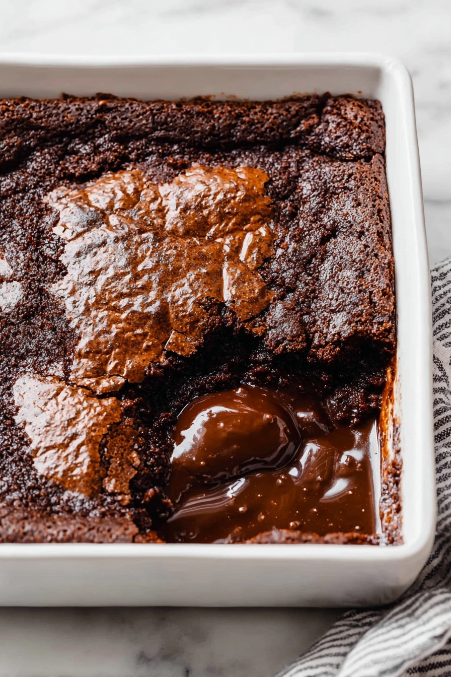 Easy Hot Fudge Cake, hot fudge cake, chocolate self-saucing cake, quick chocolate dessert, cozy chocolate cake - A square white ceramic dish holds a rich chocolate dessert with a cracked, shiny top layer that is dark brown and slightly rough in texture. One section near the front corner is scooped out, revealing a thick, glossy, melted chocolate layer beneath the top crust. The melted chocolate is smooth and dark, contrasting with the more solid and matte top layer. The dish sits on a white marbled surface, and a striped cloth partially visible under one edge. photo taken with an iphone --ar 2:3 --v 7