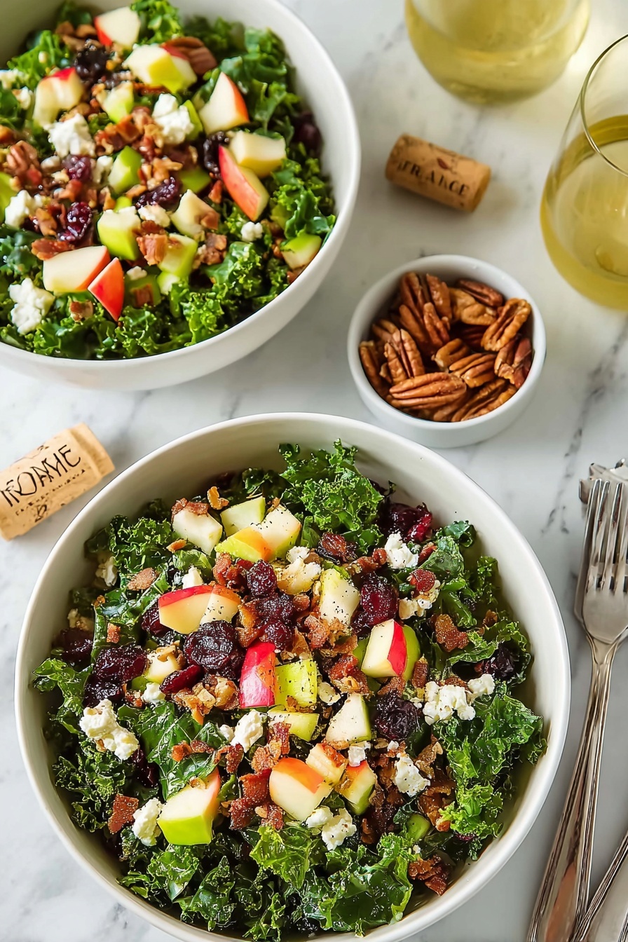 Crispy Bacon Apple Cranberry Salad, Fruit and Bacon Salad, Sweet and Savory Salad,Festive Salad with Cranberries, Easy Salad with Candied Pecans - Two white bowls filled with layered salad sit on a white marbled surface. Each bowl has a dense layer of dark green curly kale leaves at the bottom. On top of the kale are scattered pieces of red and yellow apple chunks with skin on, small light green celery bits, white crumbled cheese, dark red dried cranberries, and small brown pecan clusters. There are also small pieces of cooked bacon with a crispy texture mixed through the salad. Nearby, there is a small white bowl with extra pecans, a glass of light yellow drink, silver forks to the upper right, and a cork with the word 