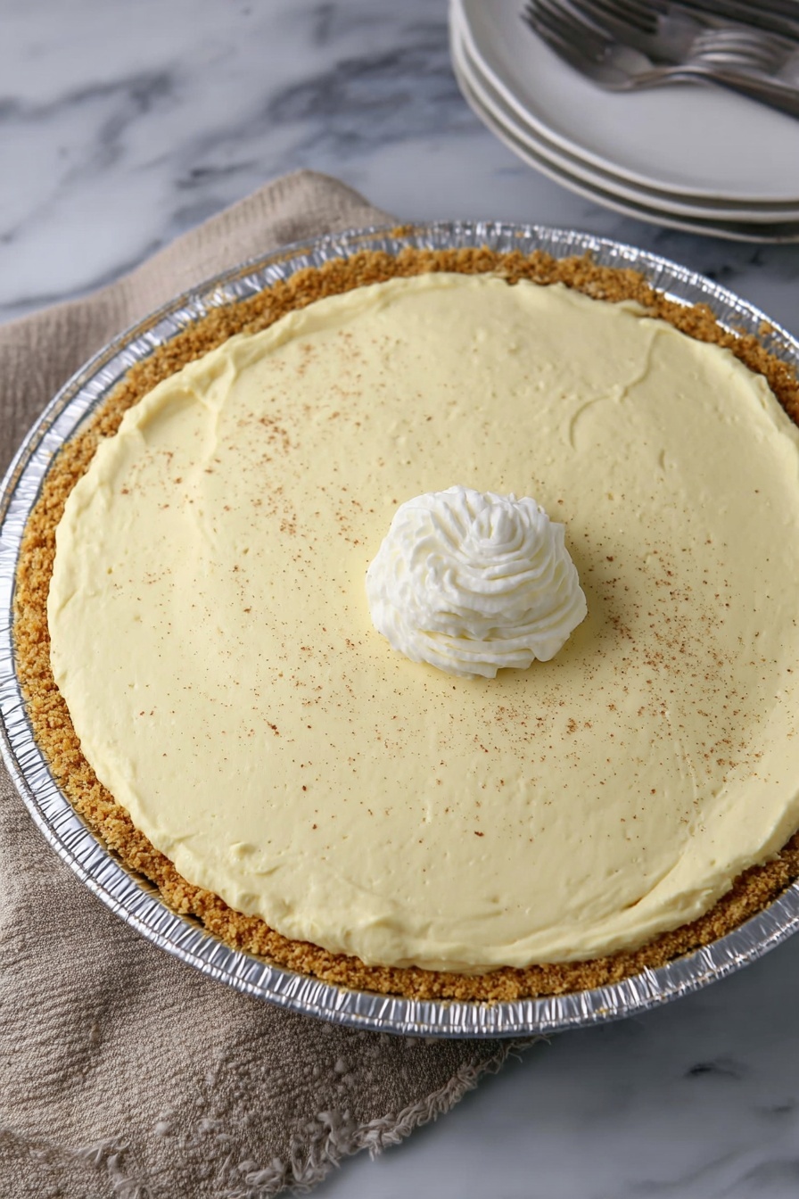 No Bake Eggnog Pie, Eggnog Pie, Holiday No Bake Desserts, Easy Festive Pies, Christmas No Bake Pie - A creamy light yellow pie with one slice missing is in a shiny round aluminum pan. It has two layers: a thick light brown crumb crust at the bottom and a thick smooth creamy yellow filling on top. The pie is topped with a small swirl of white whipped cream in the center and sprinkled with fine brown powder all over. The pie sits on a light brown woven cloth with a white marbled surface showing at the edge. On the right side, a partial view of stacked white plates with a fork is visible. photo taken with an iphone --ar 2:3 --v 7