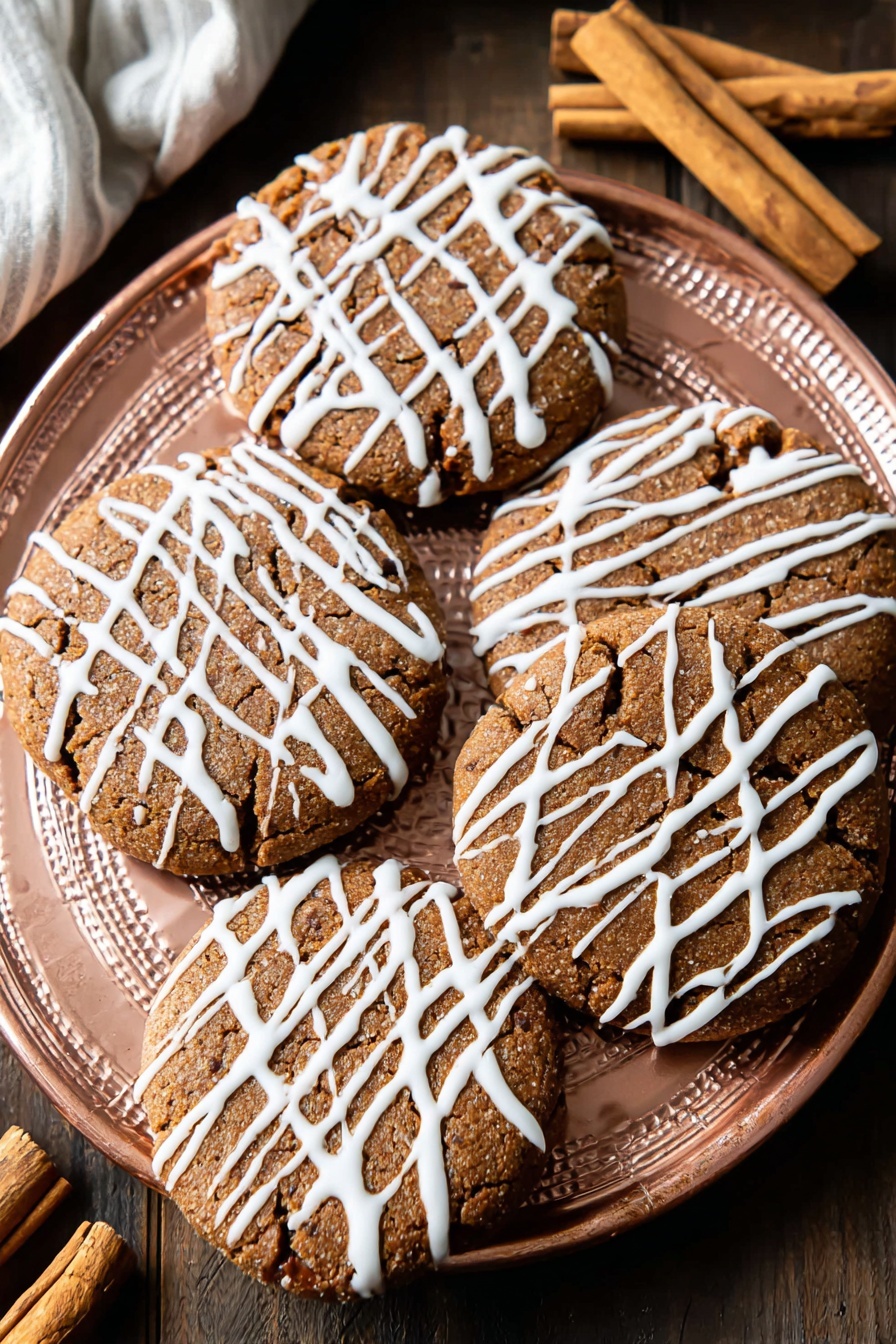 Soft Gingerbread Cookies, Gingerbread Cookies, Festive Gingerbread Cookies, Easy Gingerbread Cookies, Soft Gingerbread Cookies Recipe - A white plate holds eight round, light-brown cookies with a slightly cracked texture, each drizzled with thin white icing in a random, striped pattern across the tops. The cookies are evenly spaced on the plate, showing a soft and slightly raised surface with darker spots that hint at spices or chocolate bits. The plate sits on a white marbled surface, with a cinnamon stick and a pink cloth nearby, adding warm tones to the scene. Photo taken with an iphone --ar 2:3 --v 7
