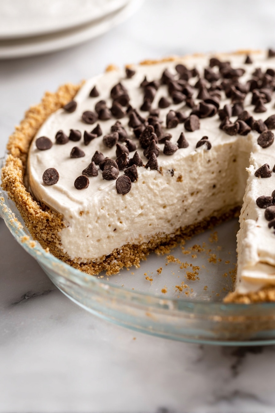 No-Bake Pistachio Cannoli Pie, pistachio dessert, easy no-bake pie, creamy pistachio dessert, Italian-inspired dessert - The image shows a pie with three visible layers. The bottom layer is a thick, crumbly crust in light brown with a rough texture around the edges. The middle layer is a thick, creamy white filling with a smooth but slightly fluffy texture. The top layer is a scattering of small, dark brown chocolate chips spread mainly on two visible slices of the pie. The pie sits in a clear glass pie dish that rests on a white marbled textured surface. A slice has been removed from the pie, showing the inside layers clearly. photo taken with an iphone --ar 2:3 --v 7