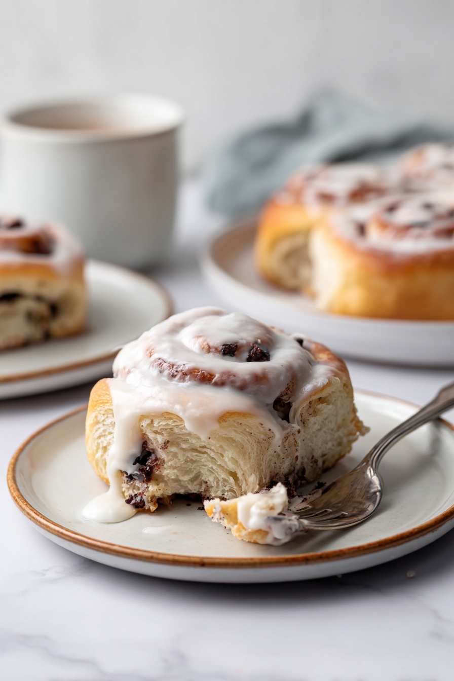 S'mores Rolls with Toasted Meringue, s'mores dessert, campfire inspired treats, chocolate graham rolls, toasted meringue dessert - The image shows a single cinnamon roll with visible layers of soft dough filled with dark chocolate chunks, placed on a white plate with a light brown rim. The top layer of the cinnamon roll is covered with white icing that drips down the sides, creating a creamy texture. Part of the roll is pulled away by a fork lying next to it, revealing the inner swirls with chocolate pieces. In the background, more cinnamon rolls on white plates and a white cup are softly blurred, all set against a white marbled surface. Photo taken with an iphone --ar 2:3 --v 7