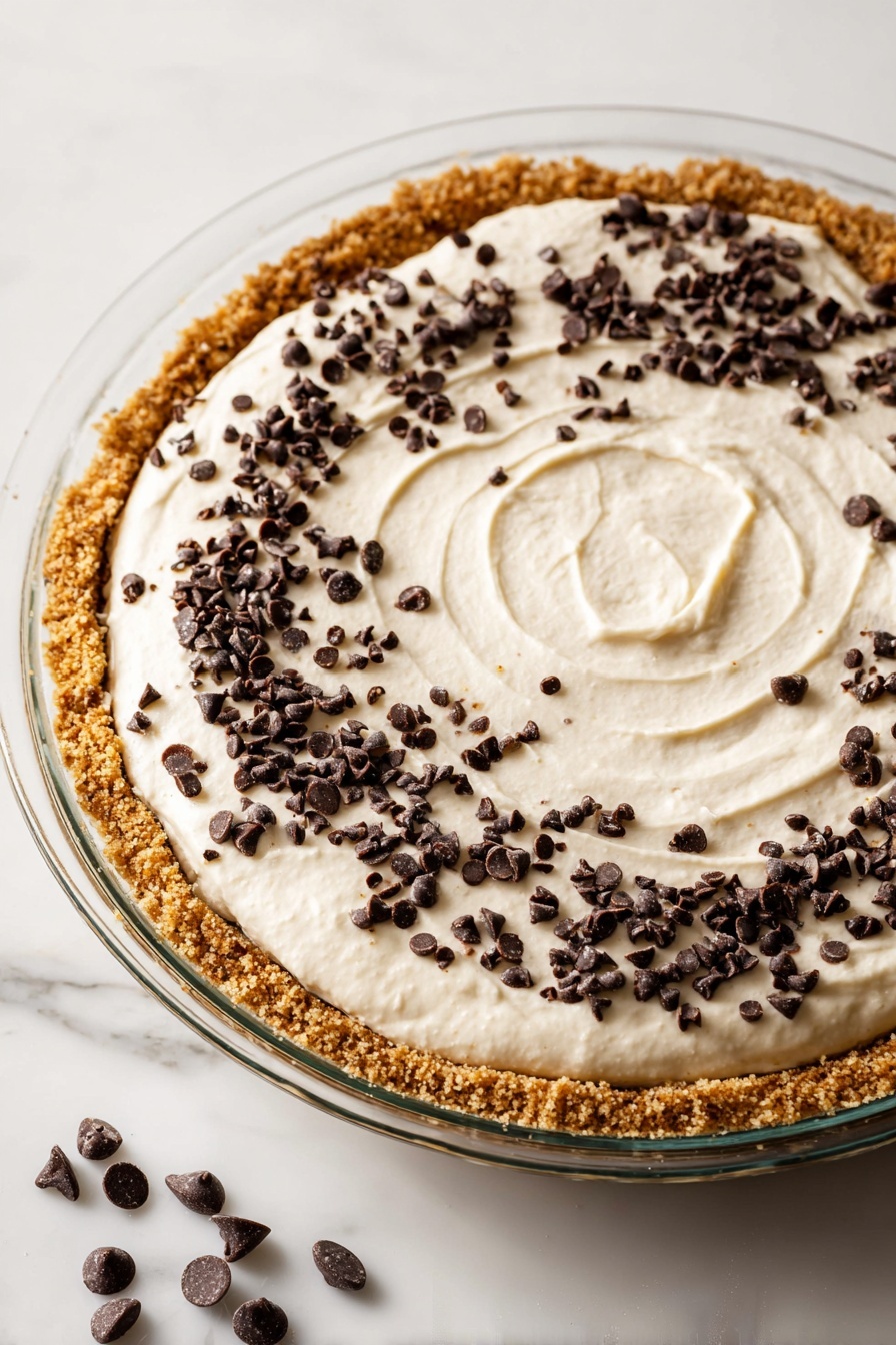 No-Bake Pistachio Cannoli Pie, pistachio dessert, easy no-bake pie, creamy pistachio dessert, Italian-inspired dessert - A glass pie dish holds a three-layer dessert placed on a white marbled surface. The bottom layer is a thick, crunchy, golden-brown crust that lines the entire base and edges of the dish. On top of the crust, there is a smooth, creamy, white filling spread evenly with a subtle spiral texture in the center. The top layer is decorated with scattered small, dark chocolate chips spread mostly near the outer edges, becoming sparser towards the center. A few more chocolate chips are loosely placed on the white marbled surface near the dish. The lighting is bright and clear, highlighting the textures well. Photo taken with an iphone --ar 2:3 --v 7