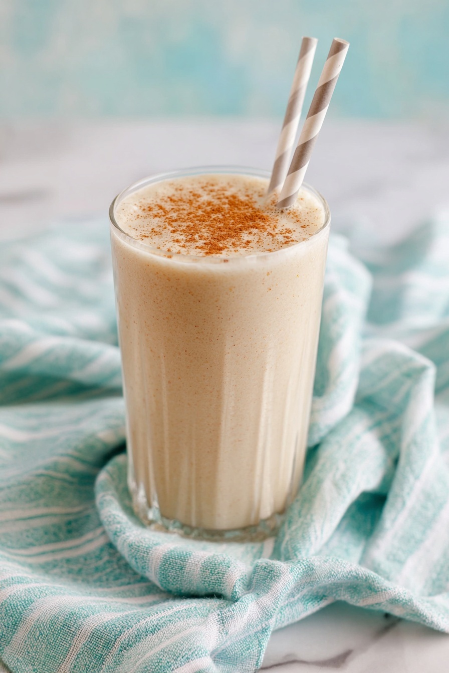 Vanilla Chai Protein Shake, healthy chai protein smoothie, quick vegan protein shake, cozy breakfast drink, easy chai protein recipe - A tall clear glass filled with a creamy, light beige drink that has small specks mixed throughout, showing a smooth and thick texture. The top of the drink is sprinkled evenly with a light brown powder. The glass sits on a green and white striped cloth on a white marbled surface. The background is soft and out of focus with light creamy tones. photo taken with an iphone --ar 2:3 --v 7